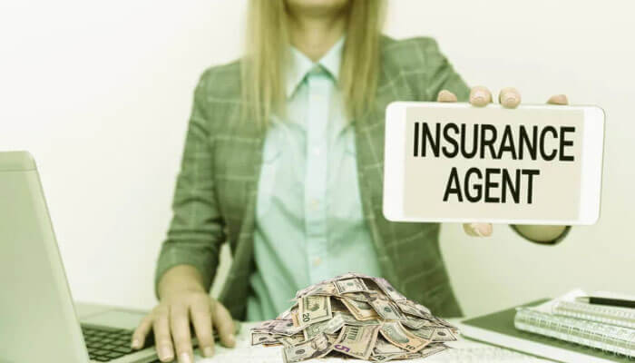 Businesswoman holding a sign that reads "Insurance Agent" with cash on the desk, symbolizing how insurance agents make money through commissions and earnings.