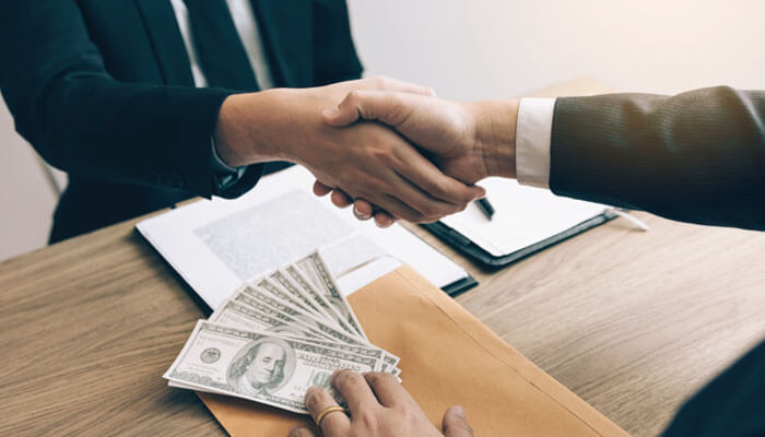 Two businessmen shaking hands with cash and documents on the table, representing a commission deal and illustrating how insurance agents make money.