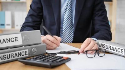 What Is An Insurance Underwriter – A professional in a suit analyzing insurance documents at a desk with binders labeled Insurance, calculator, and eyeglasses.