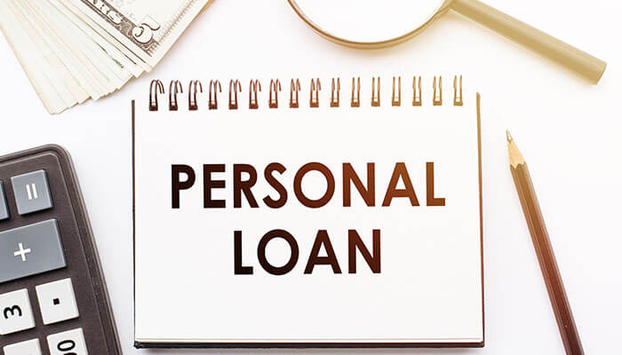Notebook with the words Personal Loan written on it, surrounded by calculator, money, magnifying glass, and pencil representing loan planning.