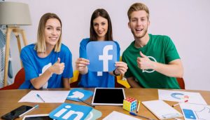 Three smiling young professionals sitting at a desk holding a large Facebook logo and giving thumbs up, representing teamwork and success in learning how to create a Facebook business page.