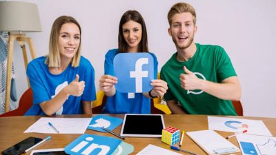 Three smiling young professionals sitting at a desk holding a large Facebook logo and giving thumbs up, representing teamwork and success in learning how to create a Facebook business page.