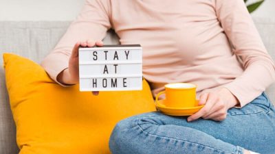 How Honey Home Insurance Keeps Your Home Safe Every Day? - Finsurancebiz A person sitting on a couch holding a lightbox sign that reads “Stay at Home” and a yellow coffee cup, symbolizing comfort and safety supported by Honey Home Insurance.