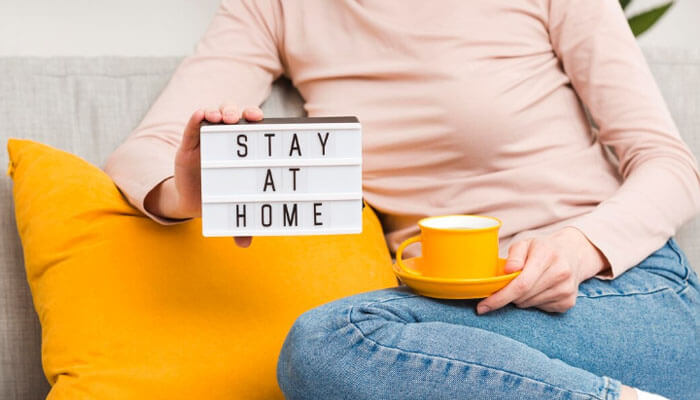 How Honey Home Insurance Keeps Your Home Safe Every Day? - Finsurancebiz A person sitting on a couch holding a lightbox sign that reads “Stay at Home” and a yellow coffee cup, symbolizing comfort and safety supported by Honey Home Insurance.