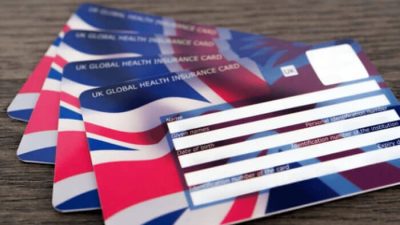 Close-up of multiple UK Global Health Insurance Card (GHIC) cards stacked on a wooden table, displaying the Union Jack design and blank information fields for cardholders.